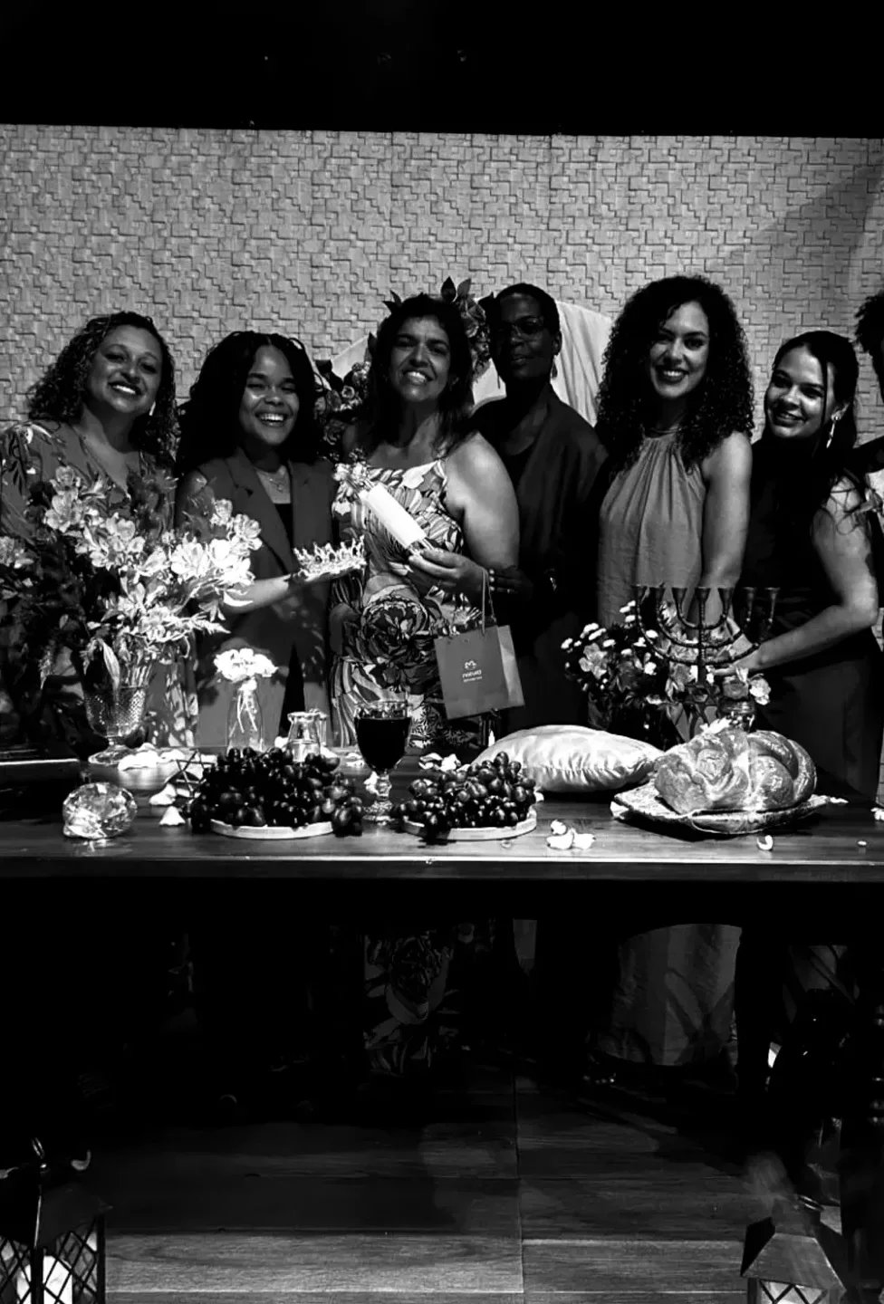 Mulheres da igreja reunidas atrás de uma mesa com flores, pães e uvas no Dia da Mulher.